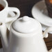 A white Homer Laughlin Seville Ivory teapot with a lid on a table with cups.