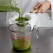 A person pouring green liquid through a Tablecraft fine tin mesh strainer into a measuring cup.