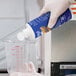 A gloved hand pours blue liquid from a blue and white container into a measuring cup.