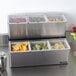 A San Jamar stainless steel condiment bar with fruit in containers.