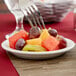 A fork holding a piece of fruit on a Tuxton Colorado narrow rim china plate.