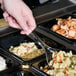 A hand holding a Visions clear plastic serving spoon over food in a tray.