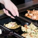 A hand holding a black Visions plastic serving spoon over a tray of food.