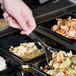 A hand holding a Visions clear plastic serving spoon over food in a tray.