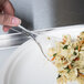 A hand holding a clear plastic serving fork over a plate of pasta.