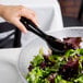 A pair of black disposable plastic tongs being used to serve salad from a bowl.