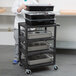 A person using a black metal Luxor serving cart to hold a metal tray of food.