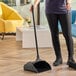 A black plastic upright dust pan with a long handle being used to collect debris from a floor.