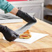 A person wearing black gloves is wrapping a sandwich with a Choice 14" x 16" insulated foil sandwich wrap sheet on a wooden countertop.