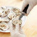 A gloved hand uses a Tablecraft Frenchman Style FirmGrip Handheld Oyster Knife to open oysters.