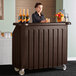 A woman standing behind a Cambro dark brown plastic portable bar with bottles on it.