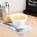 A Tuxton white china bouillon cup filled with soup on a napkin next to a bread roll and a spoon.