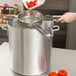 A hand uses a silver Strainer Boss Stabilizing Cradle to pour red sauce into a large pot of tomatoes.