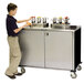 A young boy standing next to a Lakeside stainless steel condiment cart on a counter.