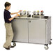 A young boy standing next to a Lakeside stainless steel condiment cart on a counter.