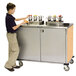 A young boy standing next to a Lakeside stainless steel condiment cart with a hard rock maple finish.