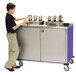 A young boy standing next to a Lakeside stainless steel condiment cart on a counter.