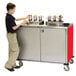 A young boy standing next to a Lakeside stainless steel condiment cart with red accents.