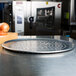 An American Metalcraft heavy weight aluminum pizza pan with holes in it sitting on a cutting board with a slice of tomato and onion.