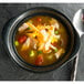 A close up of a Hall China black onion soup bowl filled with soup, chicken, and vegetables.