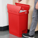 A person standing next to a red Rubbermaid Slim Jim step-on trash can with the lid open.