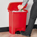 A hand putting a can into a red Rubbermaid Slim Jim step-on trash can.