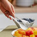 A hand holding American Metalcraft stainless steel tongs over a bowl of fruit.