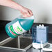 A hand pouring blue liquid from a Noble Chemical Novo soap bottle into a soap dispenser.
