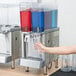 A woman's hand pressing a button on a Crathco beverage dispenser to fill a clear plastic cup with blue and red liquid.