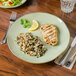 A Tuxton Artisan Sagebrush china plate with grilled chicken, rice, and salad on it with silverware and a lemon slice.