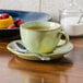 A Tuxton Artisan Sagebrush saucer with a cup of tea on it and a spoon, with a plate of raspberries.