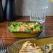 A rectangular bowl filled with salad on a table next to a glass of water.