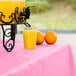 A glass of orange juice on a Creative Converting Candy Pink plastic table cover next to a drink dispenser and oranges.