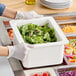 A person in gloves is putting green salad into a white Cambro food pan on a counter.