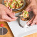 A person using an Ateco round stainless steel mold to place food in a bowl with a spoon.