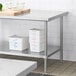 A Regency stainless steel adjustable undershelf on a white table holding white plastic containers with blue writing on them.