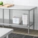 A Regency stainless steel undershelf for a work table with white containers on it.