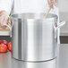 A chef holding a wooden spoon in a large silver Vollrath stock pot with vegetables.