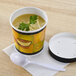 A Huhtamaki paper soup cup with a vented paper lid and a white spoon of soup on a table.