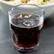 A close up of a Dinex Louis clear plastic tumbler filled with soda and ice on a table with a bowl of food.