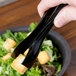 A pair of black disposable plastic tongs being used to pick up a crouton from a salad.