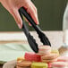 A pair of black disposable polypropylene tongs being used to pick up a macaron from a plate.