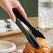 A pair of black disposable plastic tongs being used to pick up a croissant.