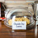 A clear glass tray with a sign and chocolate chip cookies on a table.