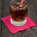 A glass with ice and a straw with a red and white striped straw on a table with a Creative Converting Hot Magenta Pink beverage napkin.