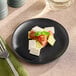 A Elite Global Solutions black melamine plate with a slice of tomato and a slice of cheese on a table with a green cloth.
