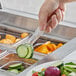 A hand using Carlisle clear polycarbonate tongs to pick up a cucumber slice from a salad.