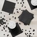 A black square Creative Converting beverage napkin on a white surface with a white container of ice cubes.