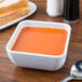 A white rounded square melamine bowl of soup on a table next to a sandwich.