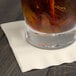 A glass of liquid with ice and a straw on an ivory Creative Converting beverage napkin.